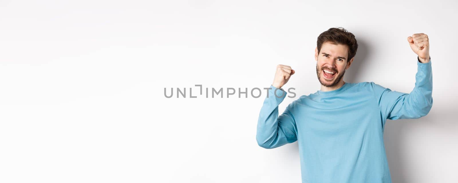 Image of happy young man triumphing, feeling like champion, winning and screaming in cheer, standing over white background.