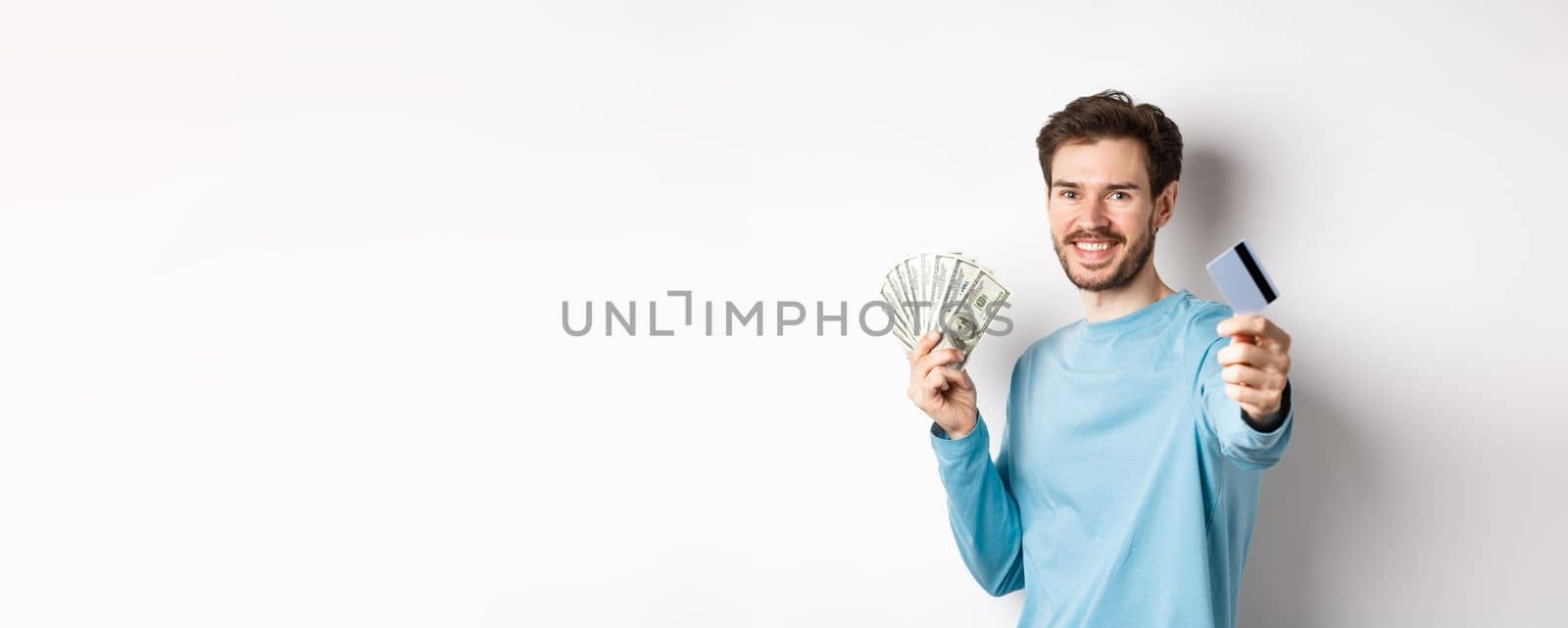 Smiling caucasian man holding money and giving you plastic credit card, standing on white background.