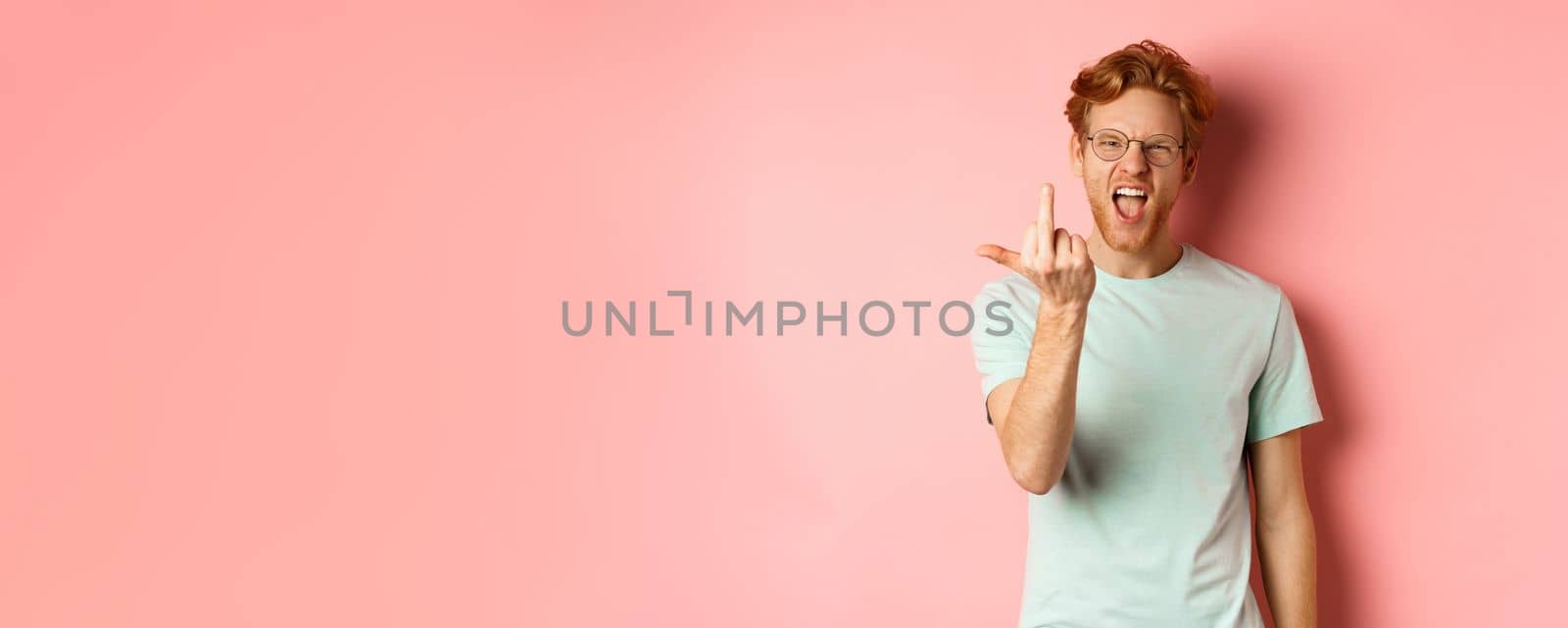 Arrogant and rude redhead man in glasses dont give a fuck, showing middle fingers at camera and frowning, standing over pink background.