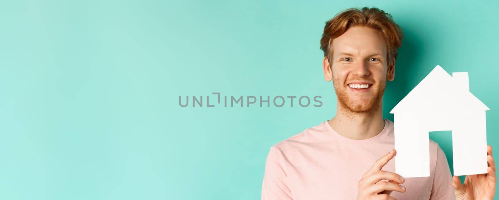 Real estate concept. Close up of redhead bearded man in t-shirt showing paper house cutout, smiling happy at camera, standing over turquoise background.