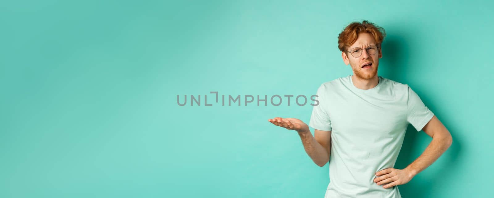 Confused redhead man trying to understand something, squinting clueless and stretch out one hand, looking puzzled, standing over mint background. Copy space