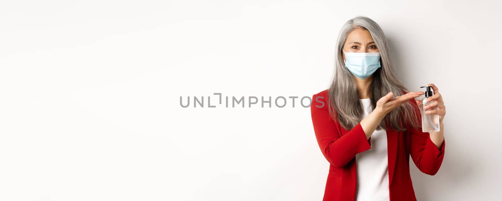 Covid, pandemic and business concept. Asian female manager in medical mask using hand sanitizer and smiling at camera, standing with antiseptic over white background by Benzoix