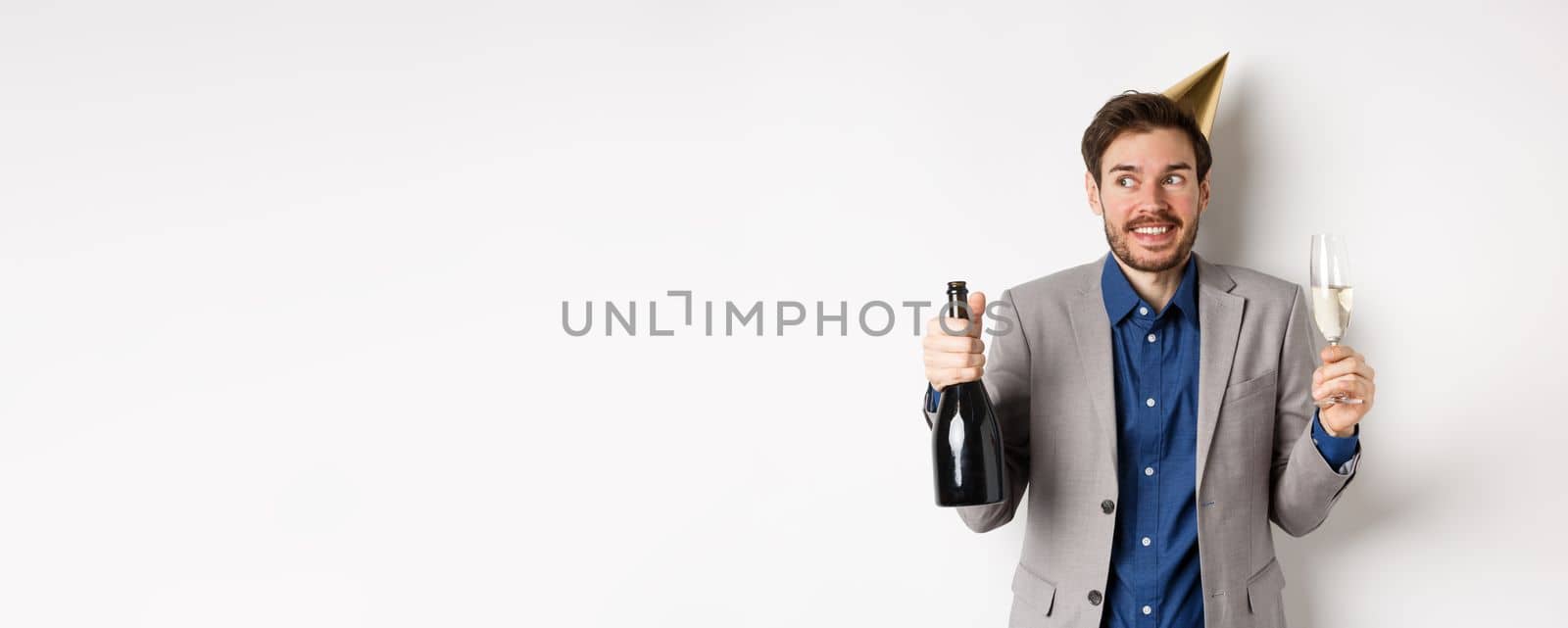 Celebration and holidays concept. Happy birthday guy in suit and party hat drinking champagne, holding bottle and glass, smiling and looking aside.