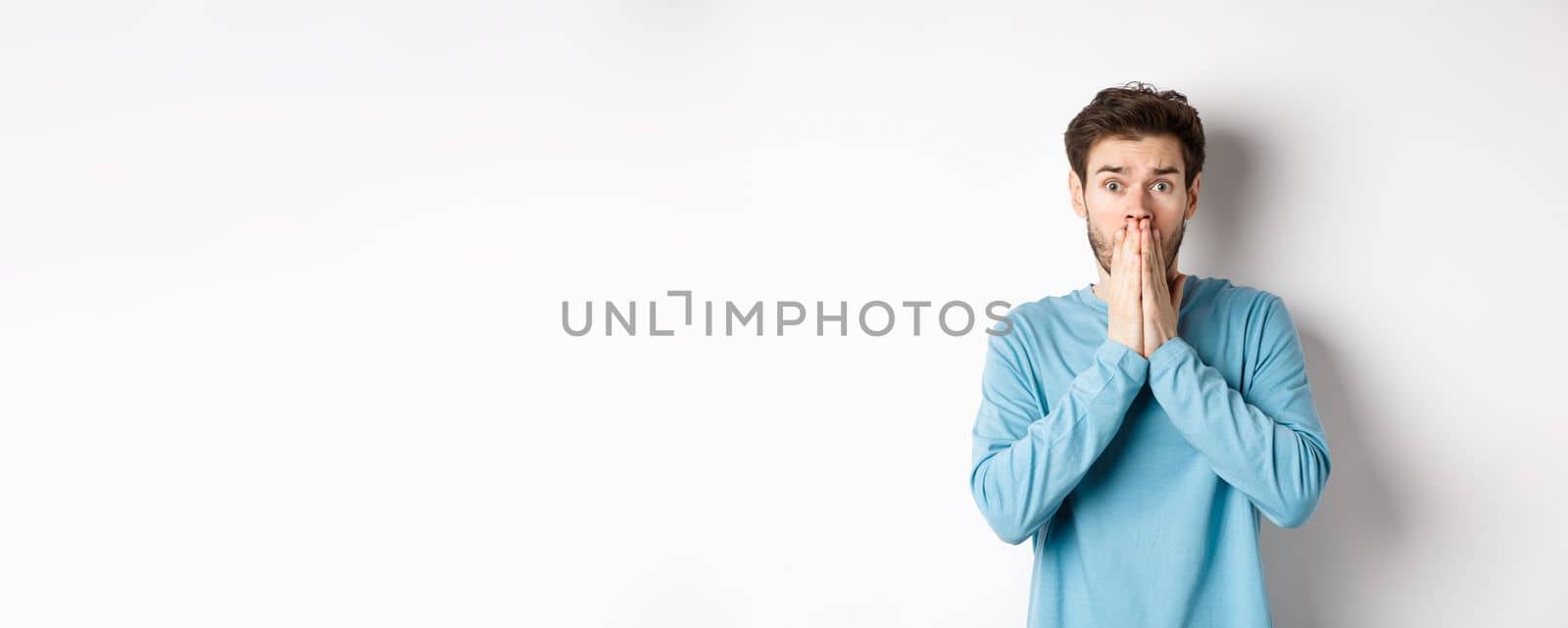 Shocked and worried young man gasping and covering mouth, look with panic at camera, standing anxious against white background.