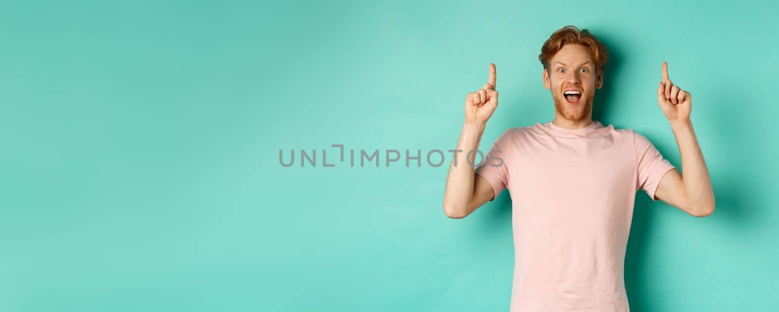 Cheerful redhead man in t-shirt pointing fingers up, staring in awe at camera and showing advertisement, standing over turquoise background by Benzoix