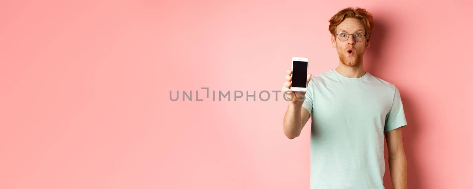 Image of handsome guy with red hair, wearing glasses and t-shirt, saying wow and showing smartphone screen, standing against pink background.