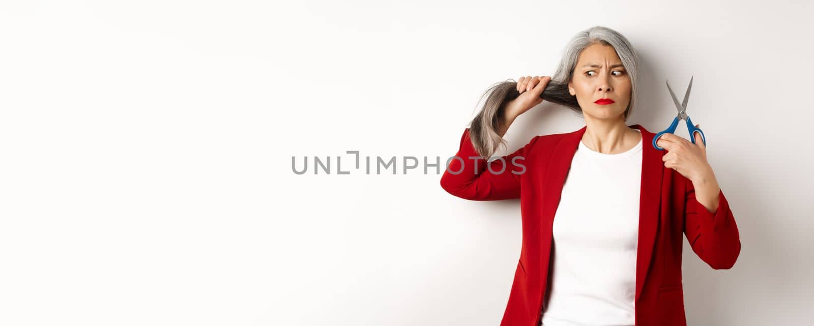 Indecisive asian woman holding scissors and looking doubtful, thinking to cut hair, changing haircut, standing over white background.