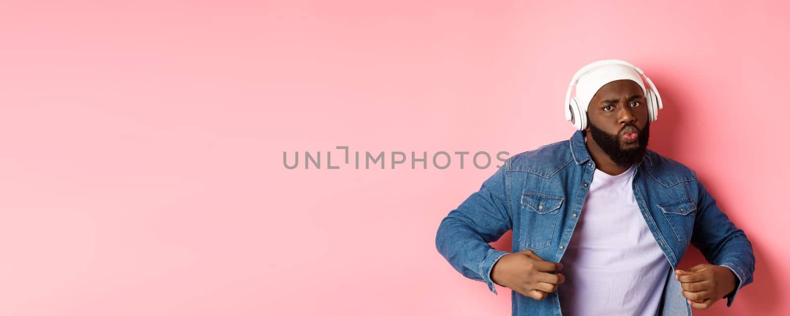 Cool and sassy Black guy dancing, listening to music in headphones, looking confident, standing over pink background by Benzoix
