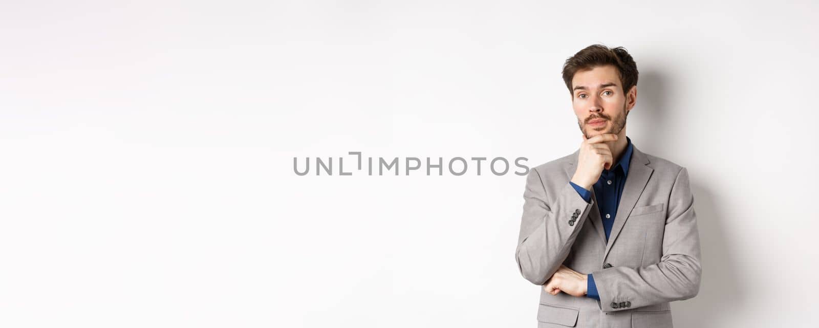 Thoughtful businessman in suit touching beard, thinking and looking at camera, deciding with pensive face, standing on white background.