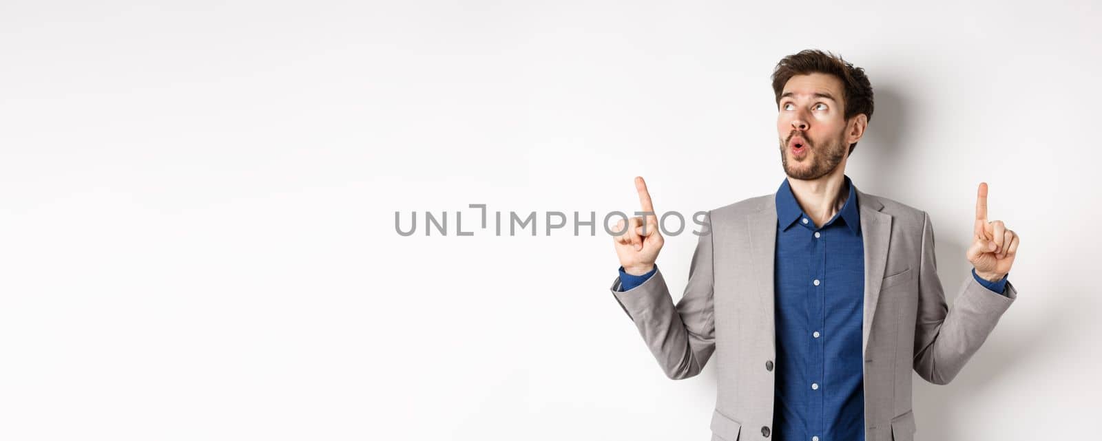 Amazed bearded guy in business suit pointing and looking up wondered, say wow and showing exciting thing, standing against white background.