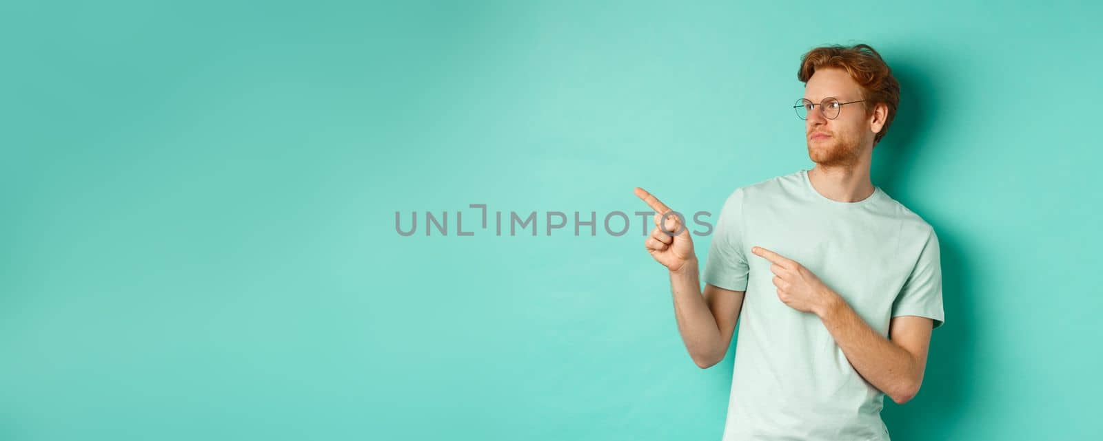 Skeptical caucasian guy with red hair and beard, wearing glasses and t-shirt, looking and pointing left disappointed, judging something bad, standing over mint background by Benzoix