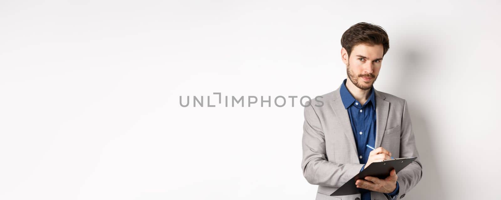 Successful businessman in grey suit taking notes, working in office clothing, holding clipboard and looking at camera, white background.