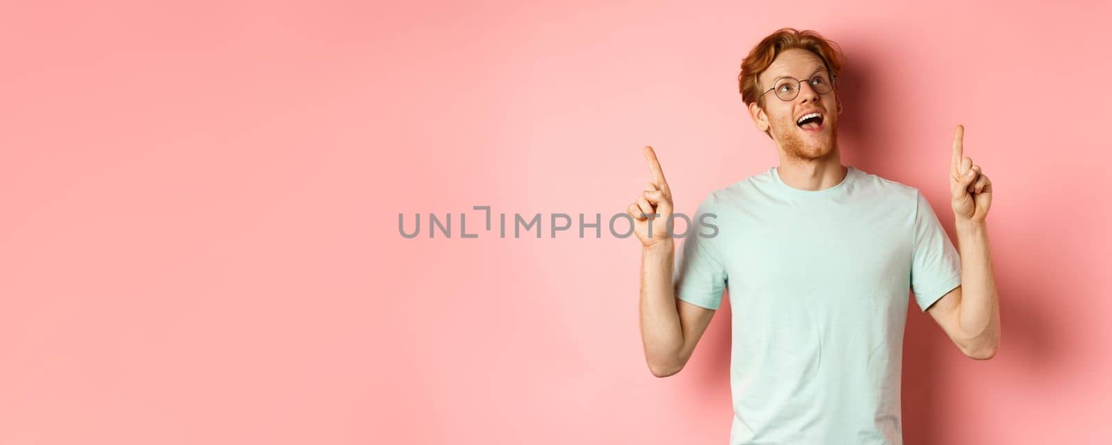 Amazed redhead guy in glasses checking out special deal, looking and pointing fingers up with amazed face, standing over pink background by Benzoix
