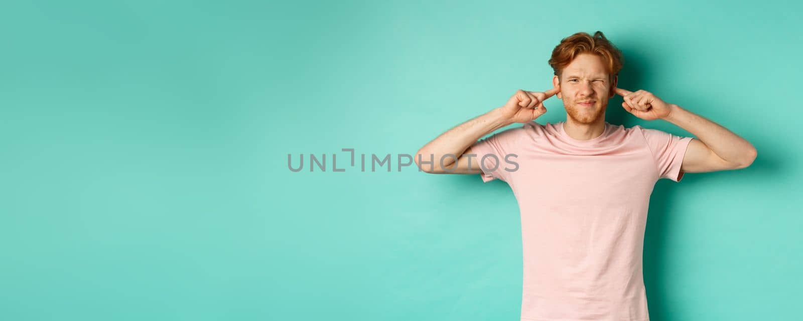 Annoyed young man with red hair and beard shut his ears and grimacing, disturbed by loud bothering sound, noisy neighbours, standing over turquoise background by Benzoix