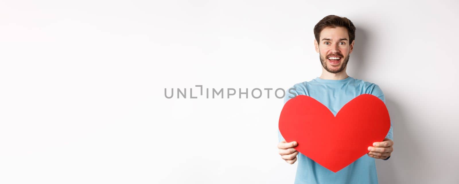 Relationship and people concept. Handsome man give valentines heart to you, smiling and saying I love you at camera, standing over white background by Benzoix