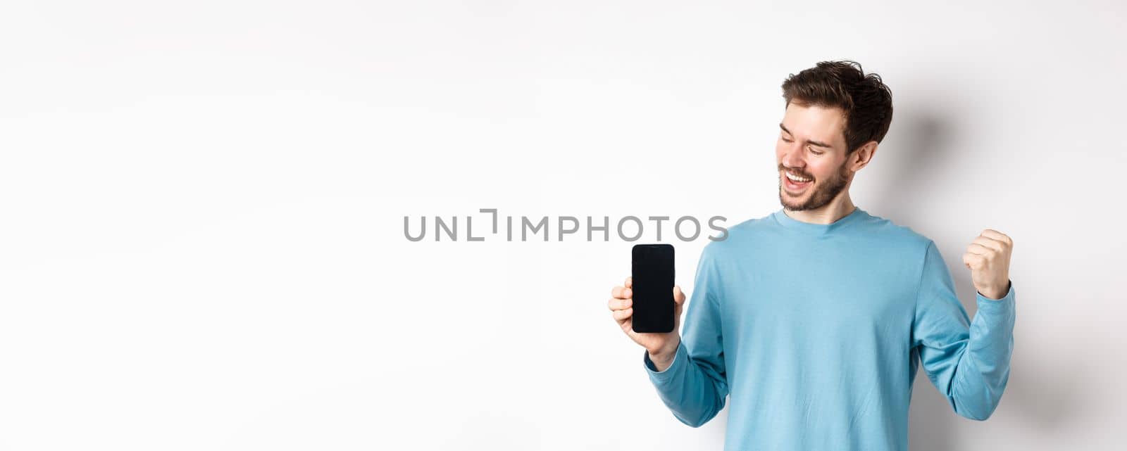 E-commerce and shopping concept. Lucky man winning online, showing empty smartphone screen and triumphing, saying yes with fist pump, standing over white background.