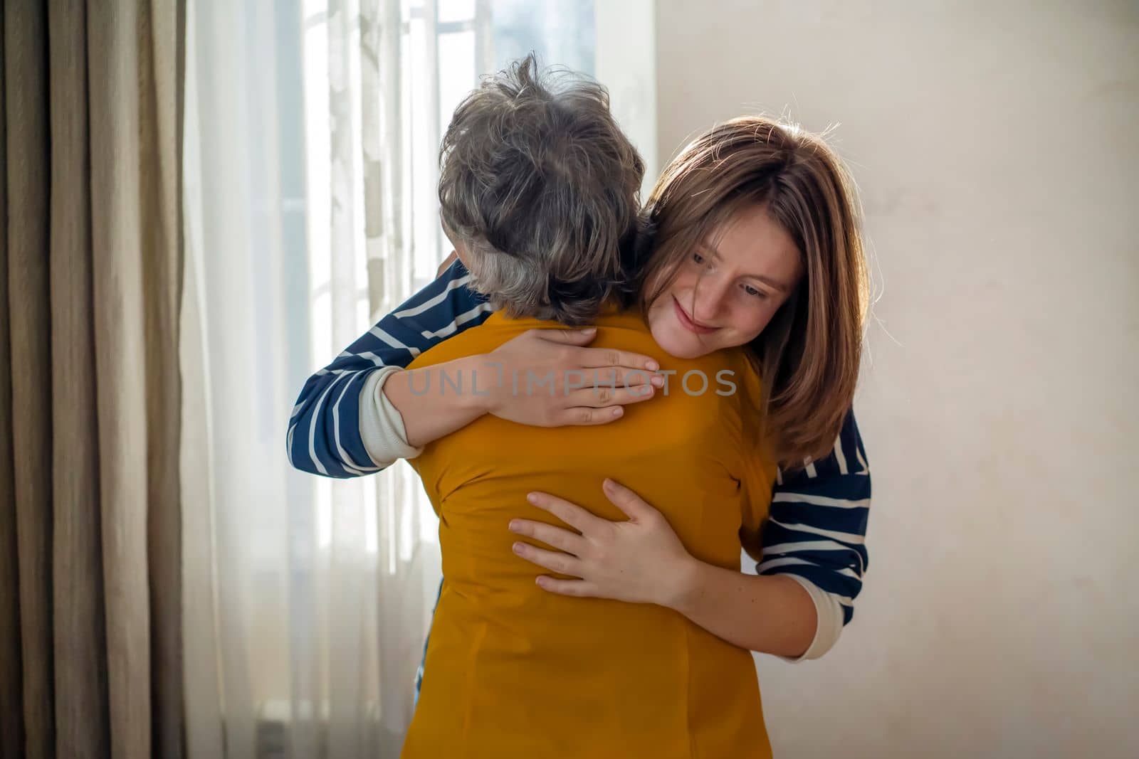 A young girl happily hugs her grandmother in a cozy house against the backdrop of the window, her daughter has a good time with her mother, lovingly cares for and supports an elderly relative.