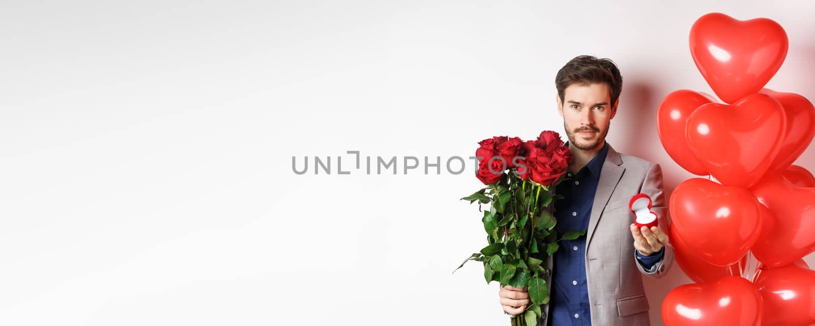 Handsome man in suit giving an engagement ring and bouquet of red roses, marry me on Valentines day, standing with heart balloons on white background.