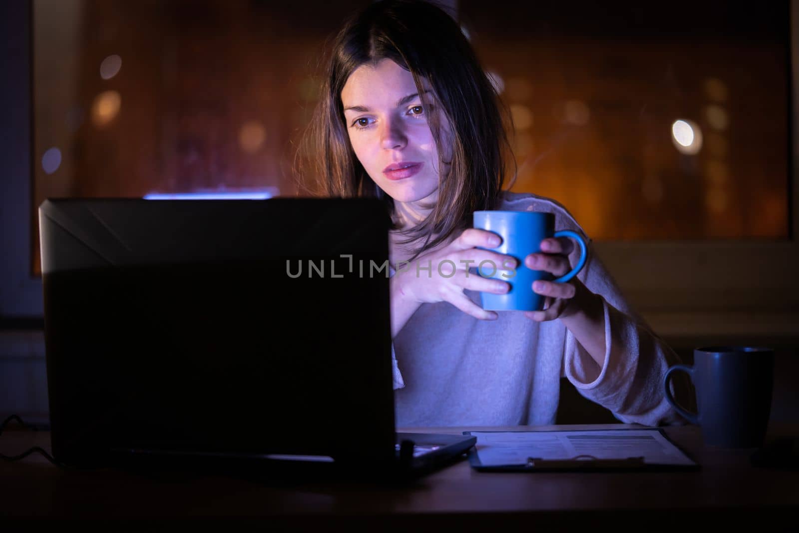 Young girl works at a laptop online at night against the background with the city lights outside the window. Woman is drinking coffee, holding a cup in her hands and reading the text on her device.