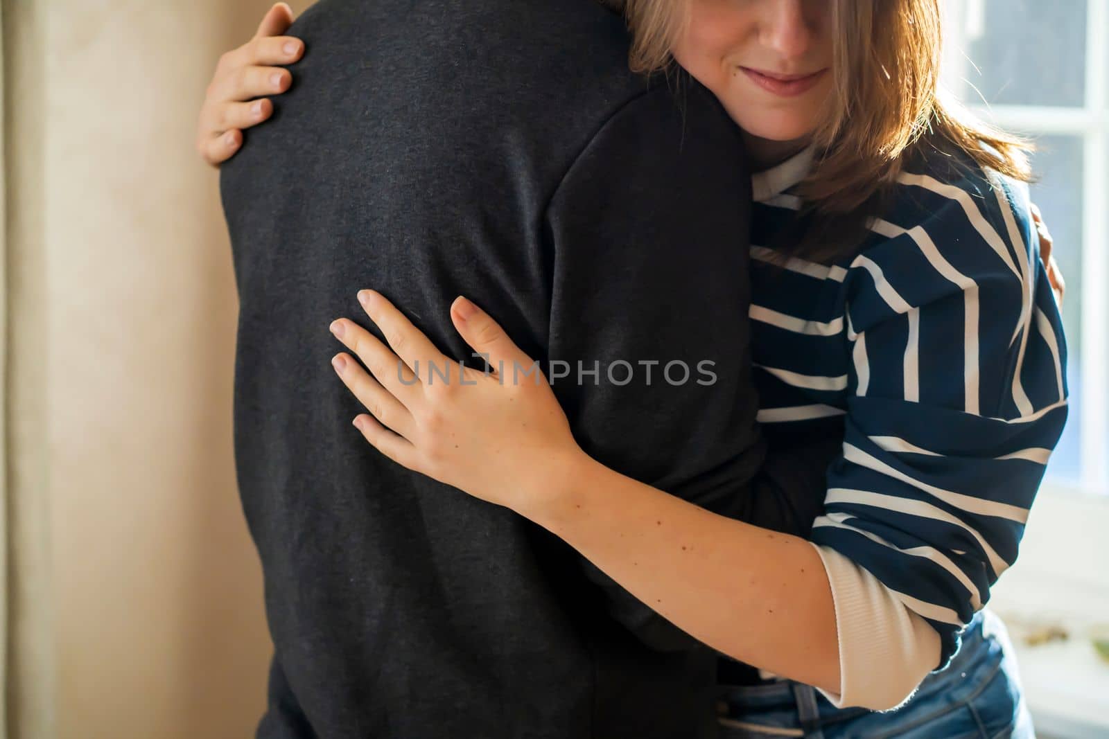 A young girl warmly and tenderly hugs her boyfriend, a couple stands against the background of a window on a sunny day, a woman begins romantic relationship. Husband and wife feel united, in harmony.