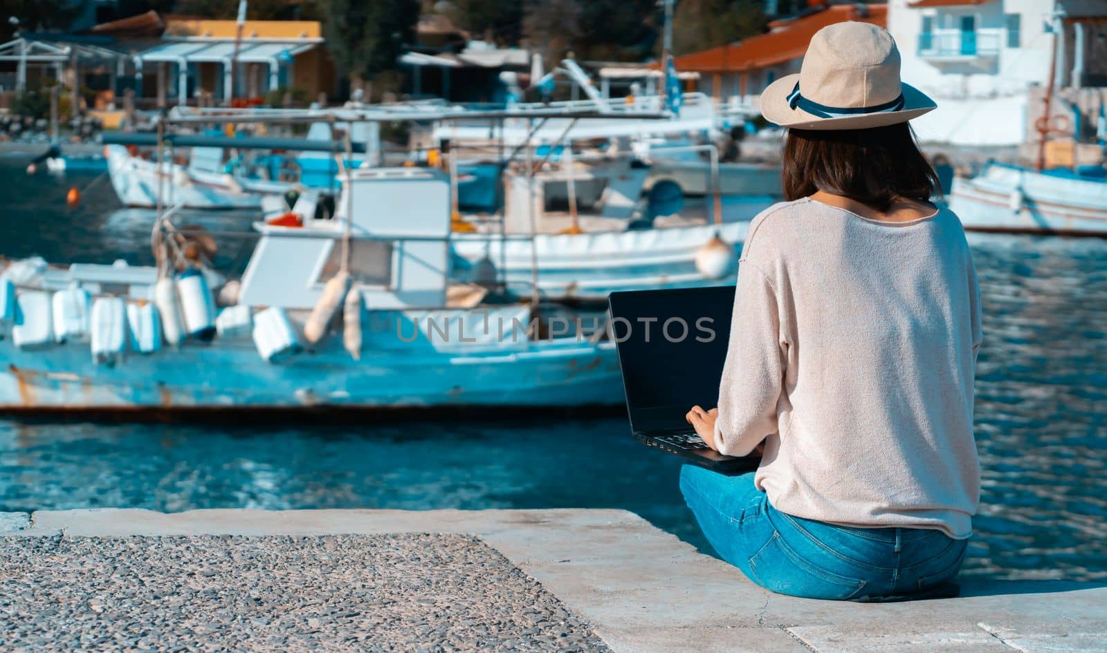 A young girl in a hat is sitting on the pier and working, typing on a laptop keyboard on a sunny day against a beautiful background of a seascape with moored yachts in the bay. Woman works and travels