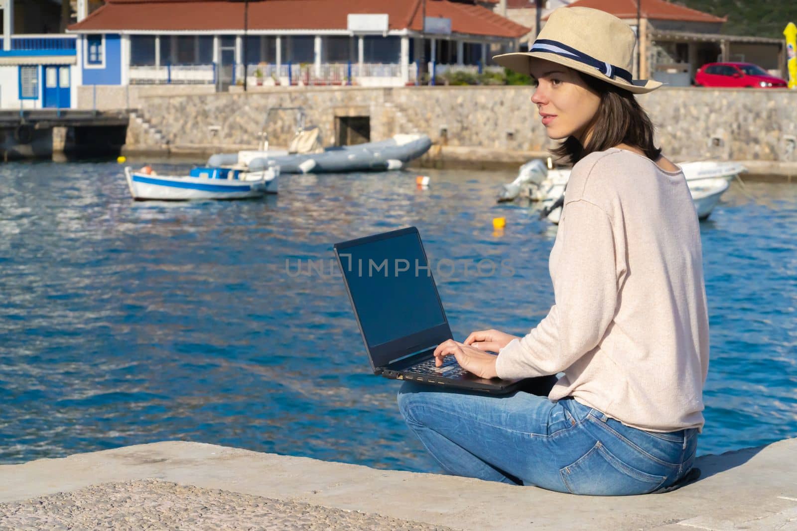 A young girl in a hat is sitting on the pier and working, typing on a laptop keyboard on a sunny day against a beautiful background of a seascape with moored yachts in the bay. Woman works and travels