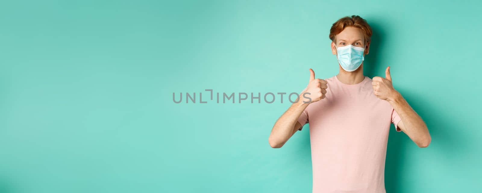 Covid-19, pandemic and lifestyle concept. Cheerful redhead guy in medical mask showing thumbs up in approval, like and praise product, standing over turquoise background by Benzoix