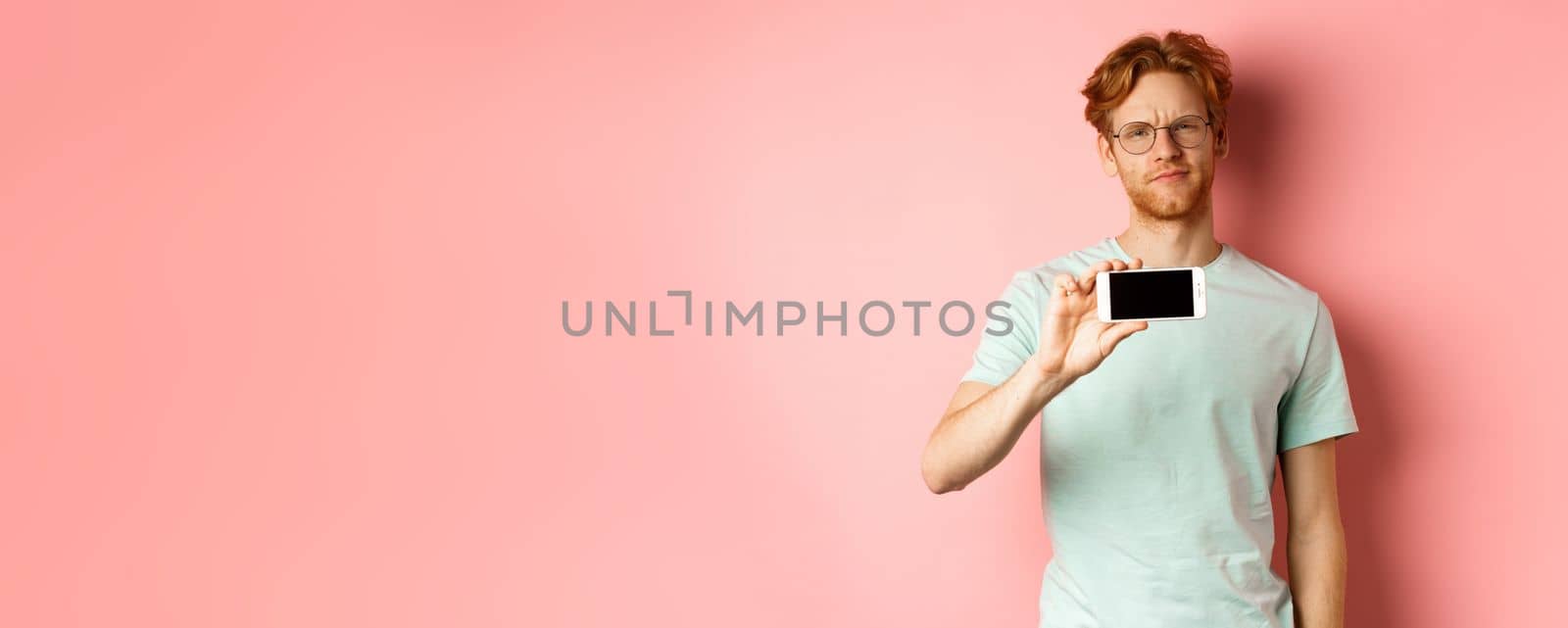 Skeptical redhead guy in glasses showing smartphone screen horizontally, smirk and frowning disappointed, standing over pink background by Benzoix