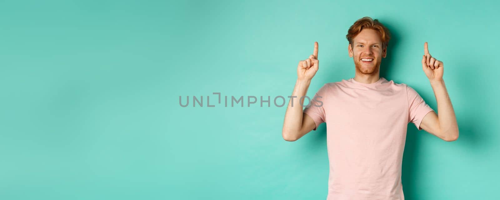 Handsome caucasian man with white teeth, smiling happy and pointing fingers up, showing promo offer, standing over mint background.