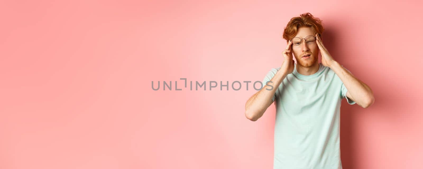 Portrait of redhead man in crooked glasses touching head and feeling dizzy or nauseous, having hangover or headache, standing over pink background by Benzoix
