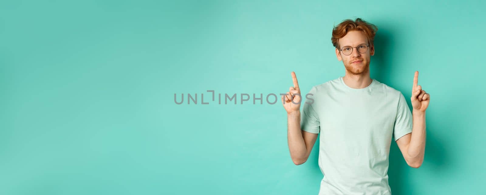 Skeptical and unamused redhead man in glasses and t-shirt, pointing fingers up and showing something useless, standing against mint background.