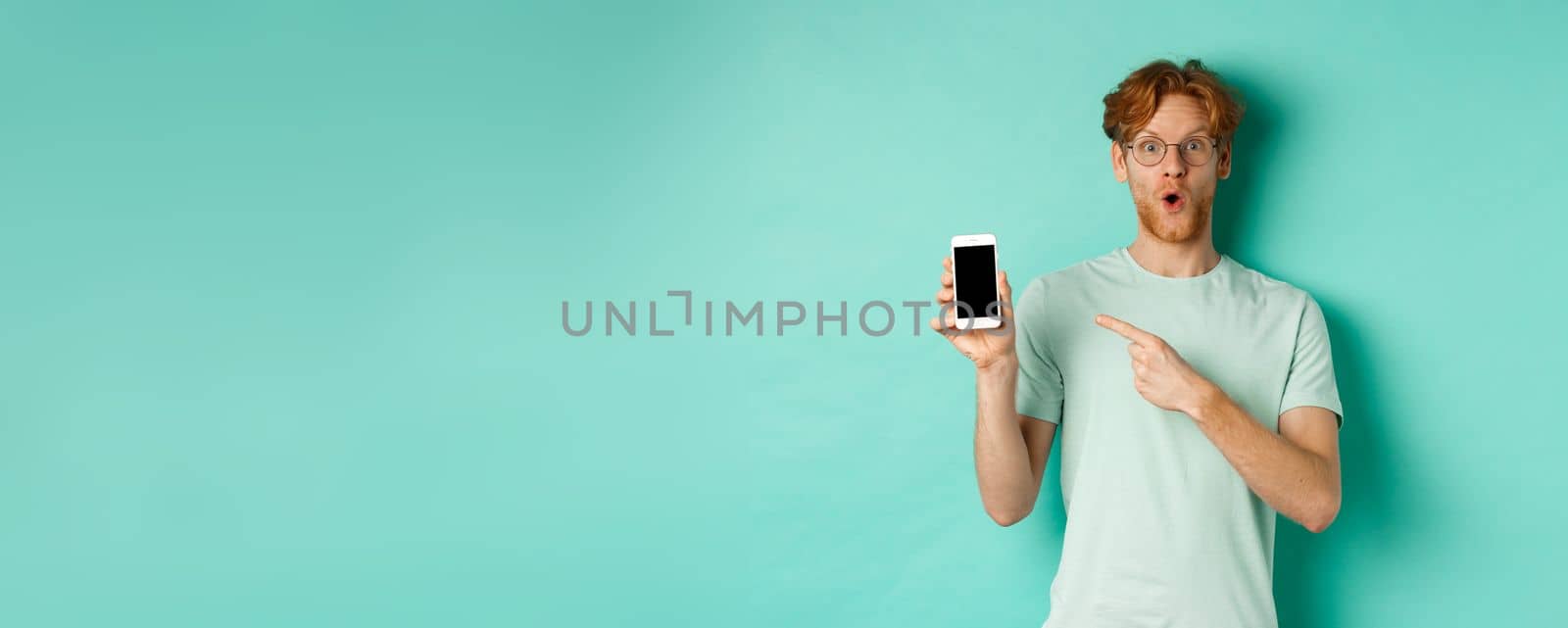 Check this out. Handsome redhead guy in glasses pointing finger at blank smartphone screen, showing online promotion, standing amazed over turquoise background.