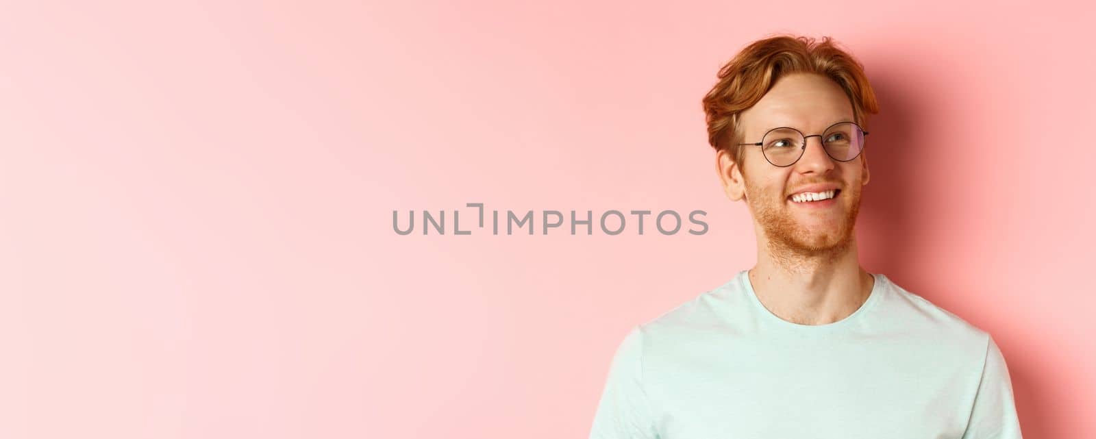 Face of handsome redhead man in glasses smiling pleased, looking at upper right corner, standing over pink background.
