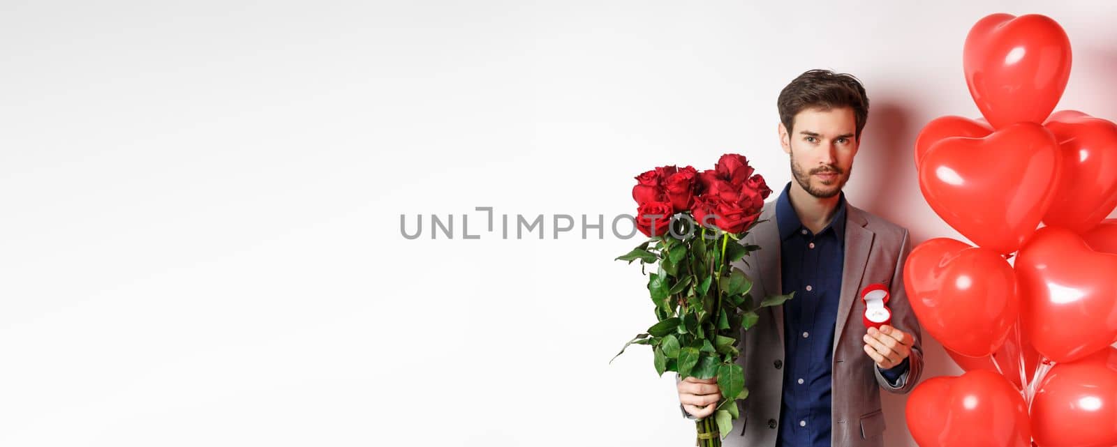 Handsome young man prepare to make proposal, holding engagement ring with bouquet of red roses, making surprise on Valentines day, standing near heart balloons, white background.