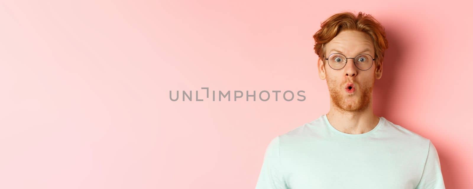 Close up of impressed redhead man in glasses saying wow, raising eyebrows surprised and staring at camera, standing over pink background by Benzoix