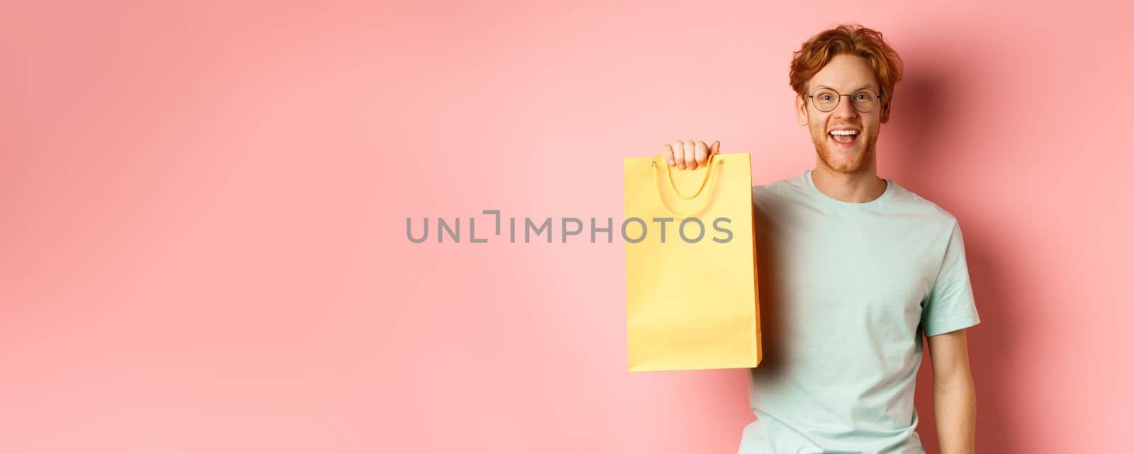 Cheerful redhead man in t-shirt, showing shopping bag and smiling, standing over pink background.