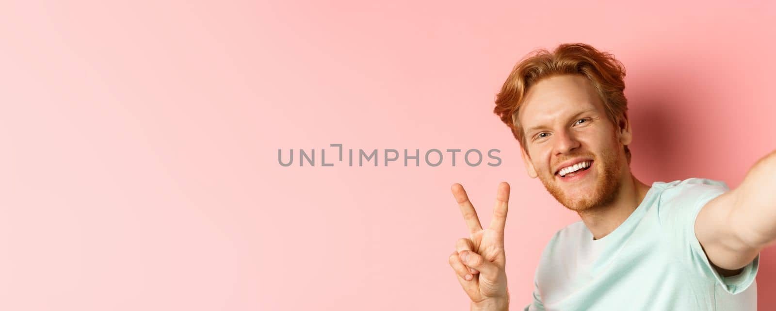 Close-up of happy redhead man taking selfie on smartphone and showing peace sign, view from phone camera, pink background by Benzoix
