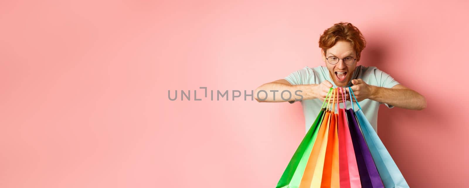 Excited young man, shopper holding shopping bags and smiling happy, looking excited at purchased items, standing over pink background.