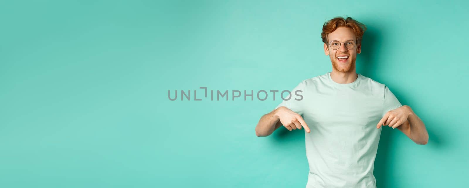 Happy young man with red hair, wearing glasses and t-shirt, showing advertisement, smiling and pointing fingers down, standing over turquoise background.