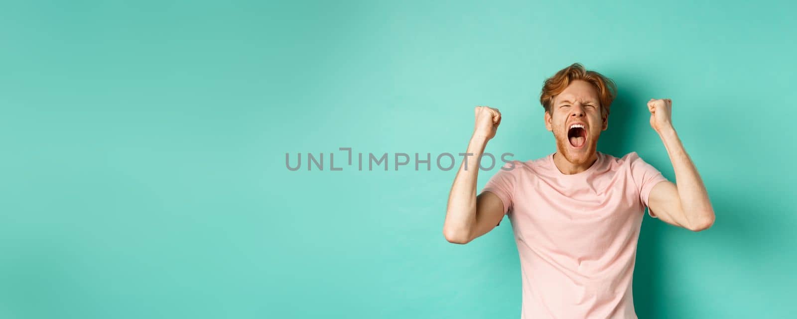 Handsome redhead man watching sports game and cheering, shouting and making fist pumps, looking at screen and watching competition, rooting for team, standing over turquoise background by Benzoix