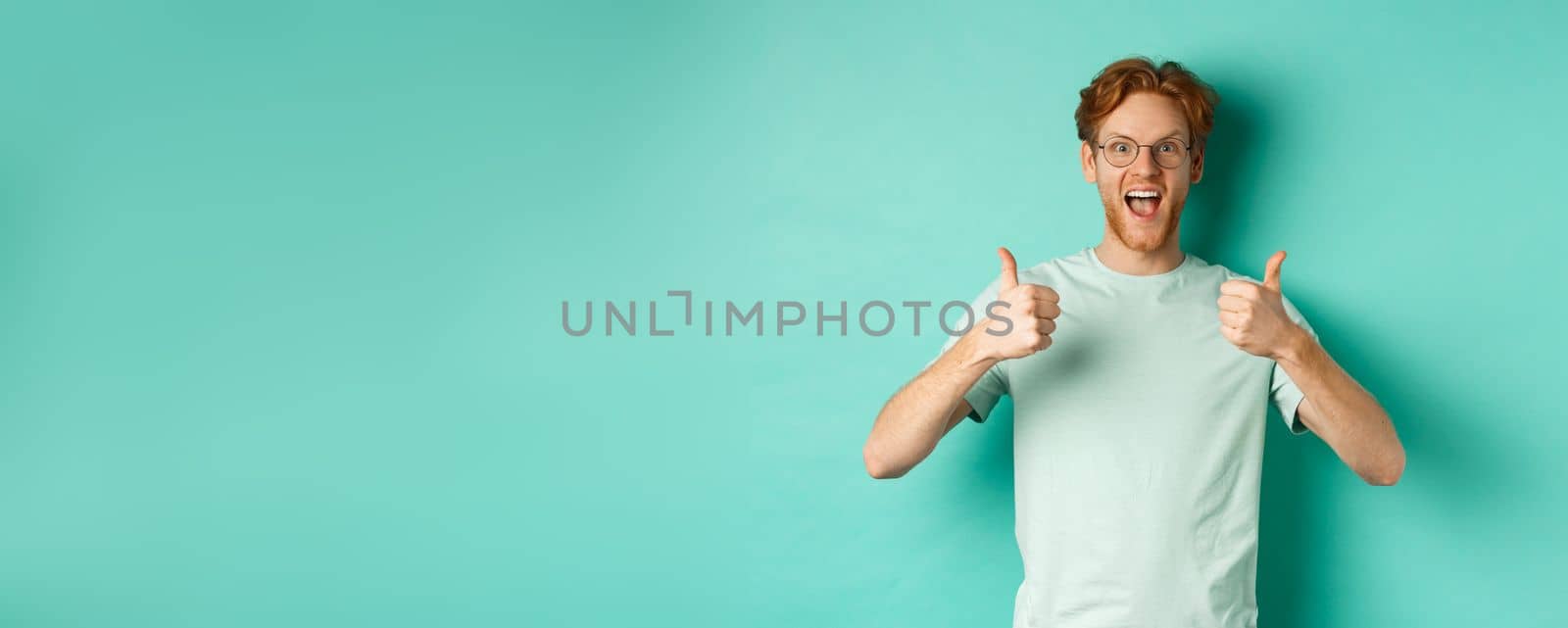 Excited young man with red hair, wearing glasses, showing thumbs-up and agree or praise something, smiling amazed and saying yes, standing over turquoise background.
