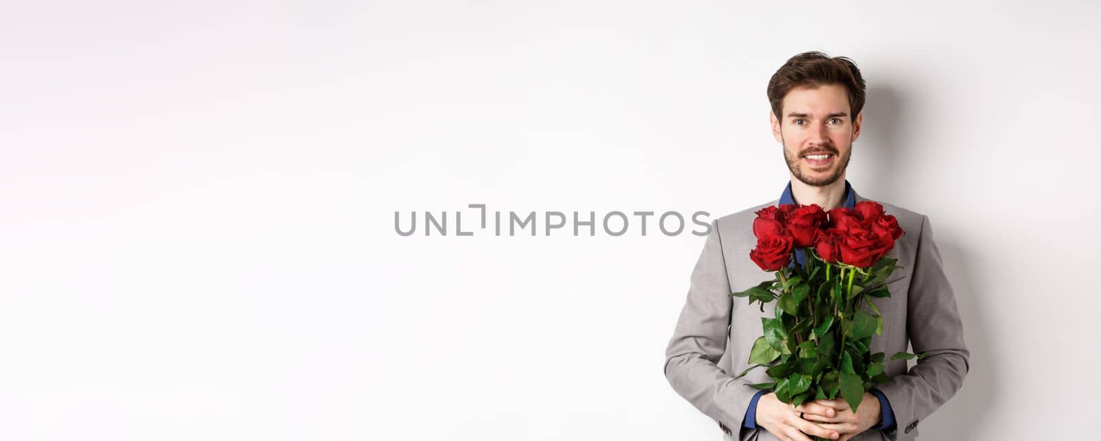 Handsome young man in suit standing with gift on valentines day, holding red roses bouquet and smiling at lover, standing over white background by Benzoix