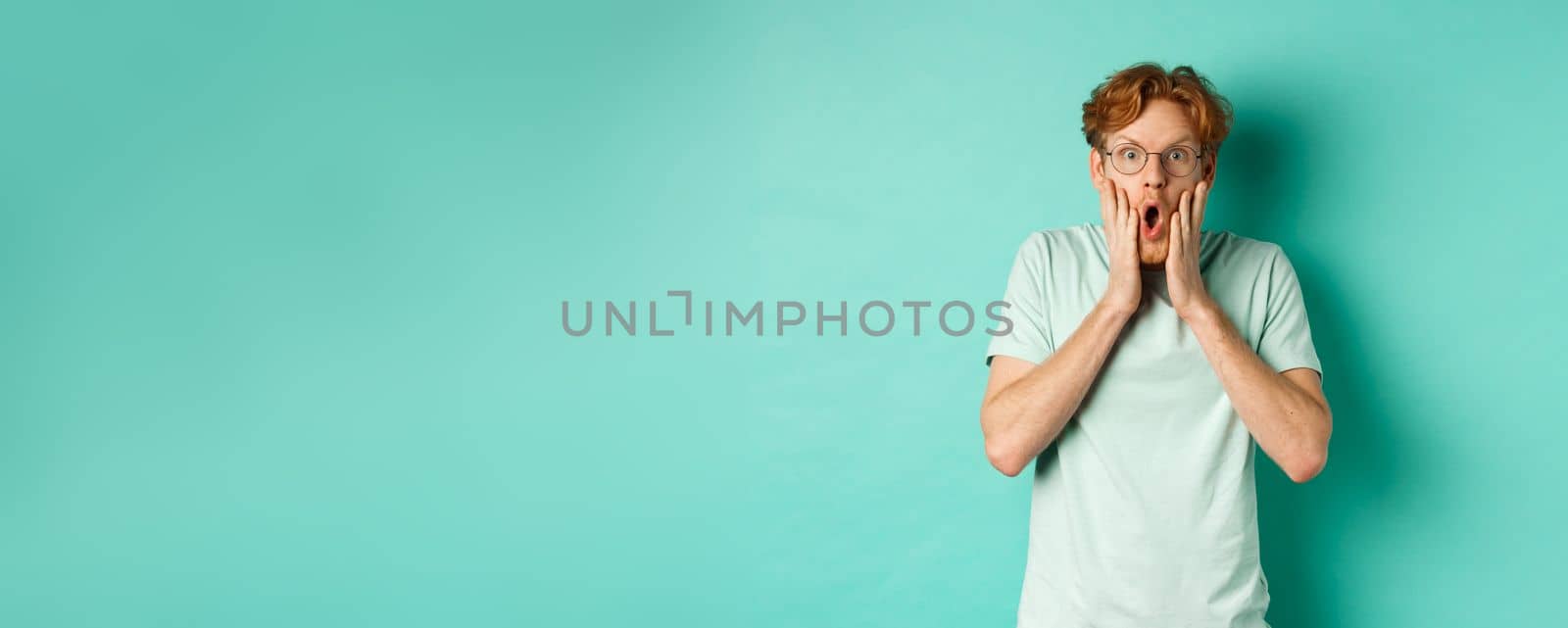 Shocked redhead guy in glasses checking out awesome promotion offer, gasping amazed and holding hands on face, staring at camera, mint background.