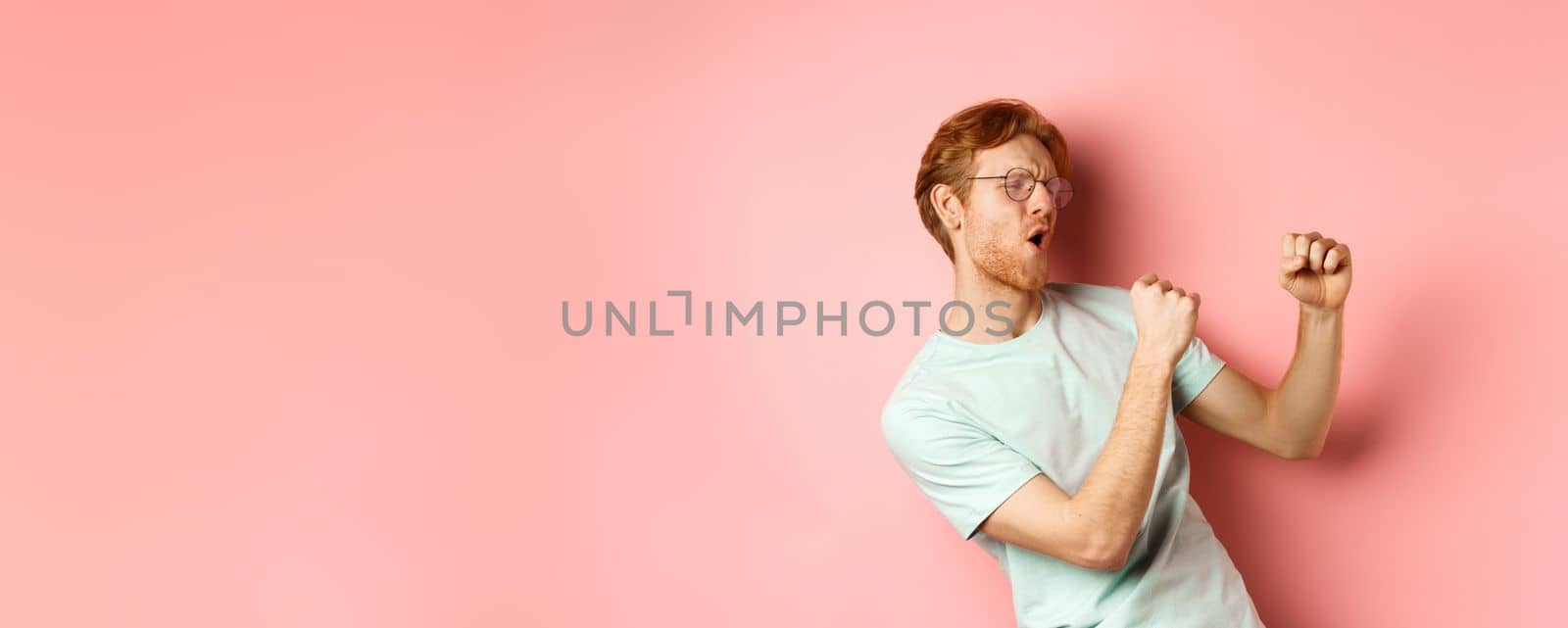 Cheerful redhead guy listening music and dancing, triumphing or celebrating success, standing over pink background.