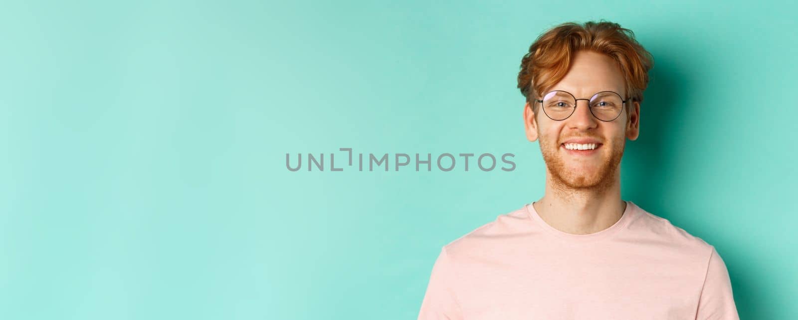 Close up of handsome redhead man in glasses looking at camera, smiling with white teeth, standing against mint background.
