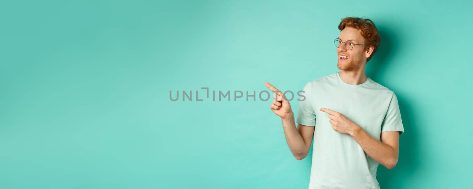 Handsome young man with red hair and beard, wearing glasses and t-shirt, pointing and looking left with amused face, checking out advertisement on copy space, mint background.