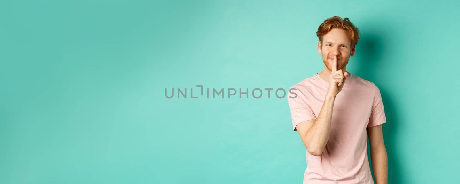 Smiling young man with red hair and beard sharing a secret, showing taboo gesture and grinning, shushing you to be quiet, standing over turquoise background by Benzoix