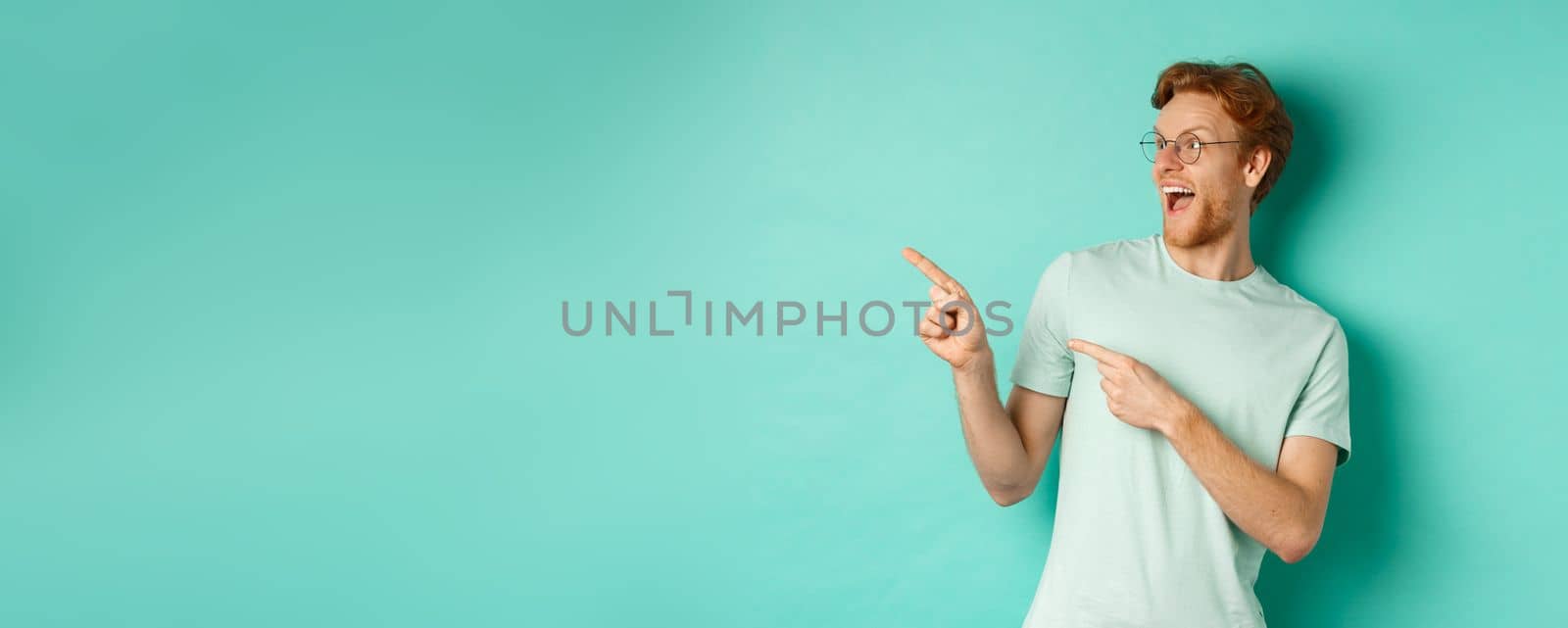 Excited young man with red hair, wearing glasses and t-shirt, pointing and looking left at awesome promotion, smiling happy at banner, turquoise background.