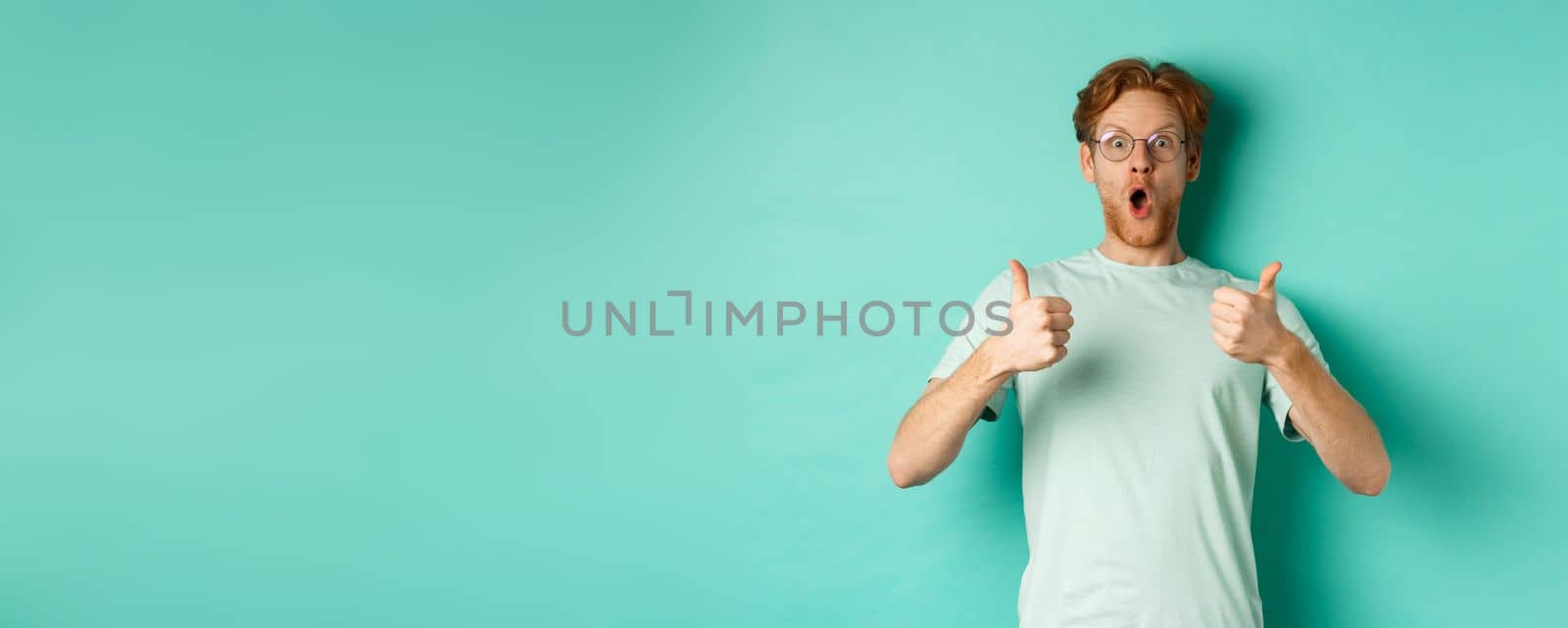 Amazed young man with red hair and beard, wearing glasses with t-shirt, showing thumbs-up and gasping in awe, checking out awesome promo offer, turquoise background.