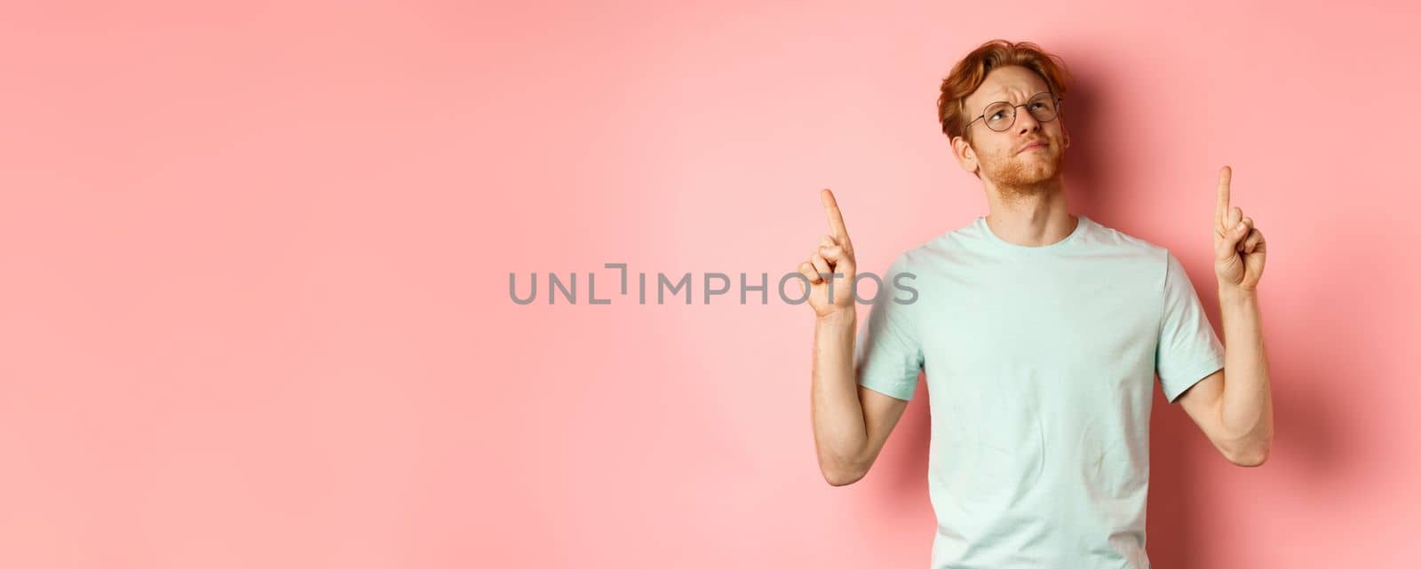 Doubtful redhead guy in glasses frowning, looking and pointing fingers up at something disappointed, standing over pink background.